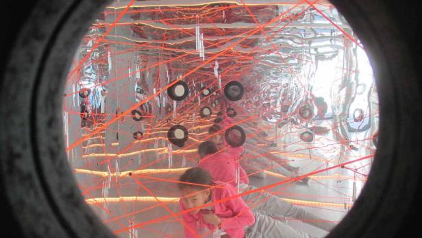 Ein Mädchen klettert durch ein Spinnennetz aus roten Laserstrahlen in einem Spiegelkabinett.