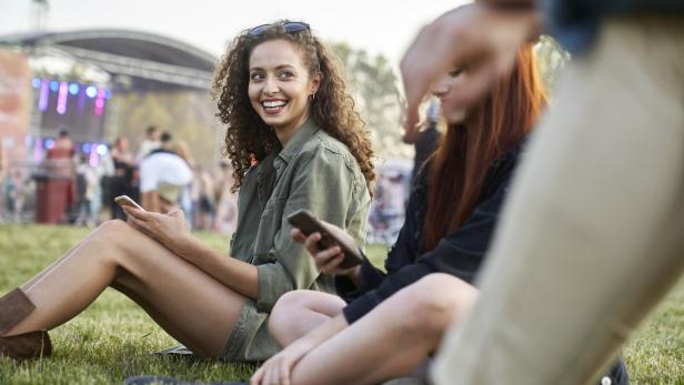 Zwei junge Frauen sitzen auf einer Wiese und nutzen ihre Smartphones. Im Hintergrund ist eine Bühne zu sehen.
