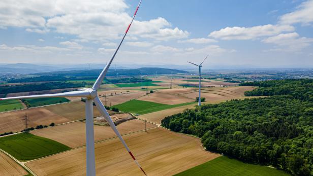 Windräder stehen auf Feldern neben einem Wald unter blauem Himmel.