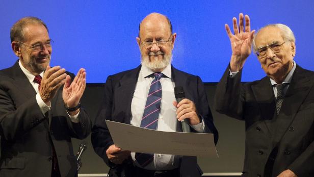 Drei ältere Männer in Anzügen stehen vor einem blauen Hintergrund, einer klatscht, einer liest, einer winkt.