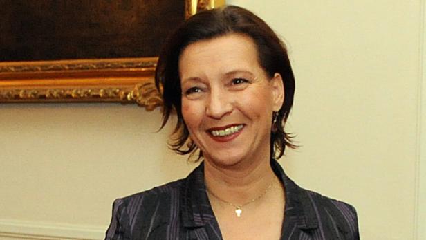 Eine lächelnde Frau mit dunklen Haaren und einem Blazer vor einem Gemälde.