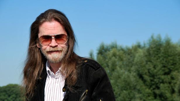 Ein Mann mit langen Haaren, Bart und Sonnenbrille vor blauem Himmel und Bäumen.