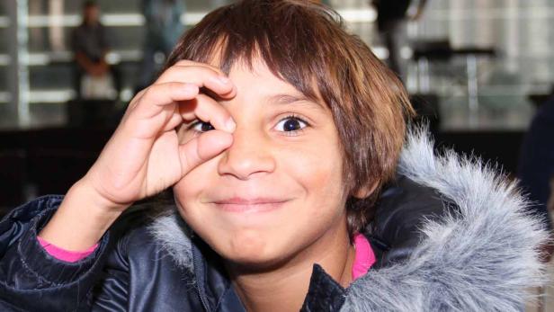 Ein lächelnder Junge mit Winterjacke formt mit seiner Hand eine Art „Ok“-Zeichen vor seinem Auge.