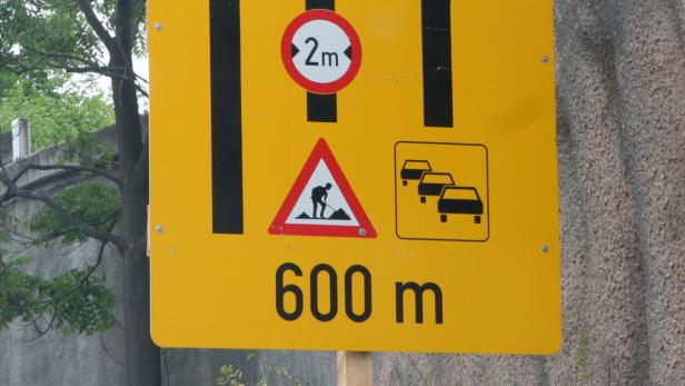 Ein gelbes Verkehrsschild warnt vor Bauarbeiten und Stau in 600 m Entfernung sowie einer Höhenbeschränkung von 2 m.