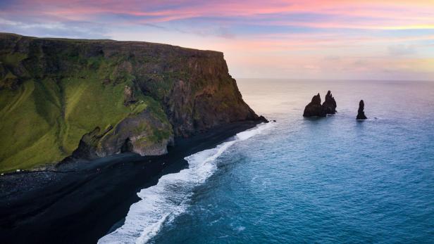 Blick auf Reynisfjara, Island, mit schwarzem Sandstrand, Basaltsäulen und Felsnadeln im Meer.