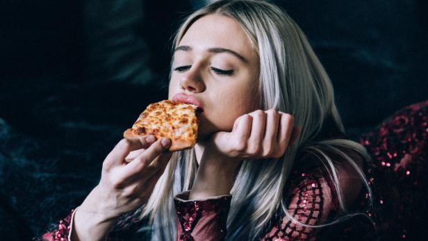 Eine Frau mit blonden Haaren isst ein Stück Pizza auf dem Bett.