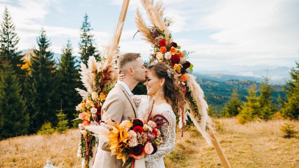 Ein Paar umarmt sich vor einem mit Blumen geschmückten Traubogen in den Bergen.