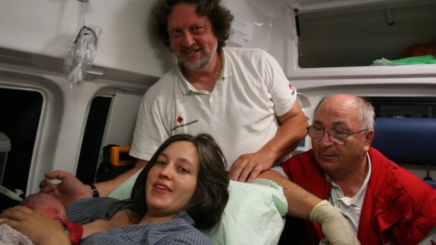 Eine Frau liegt mit ihrem Neugeborenen in einem Krankenwagen, umgeben von zwei Sanitätern.