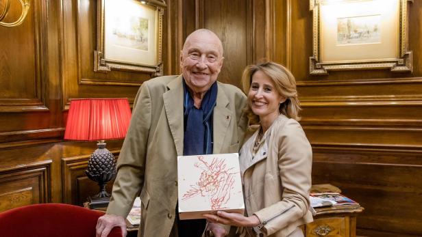 Georg Baselitz und Alexandra Winkler posieren mit einer Schachtel in einem holzgetäfelten Raum.