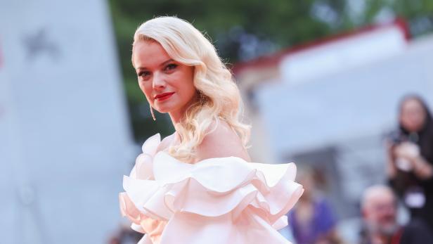 Eine Frau mit blonden, gewellten Haaren und einem rosa Kleid mit Rüschen.