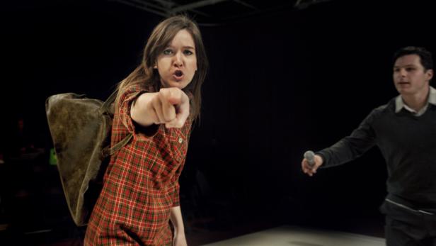 Eine Frau in karierter Kleidung zeigt mit dem Finger auf den Betrachter, während ein Mann mit Mikrofon im Hintergrund steht.