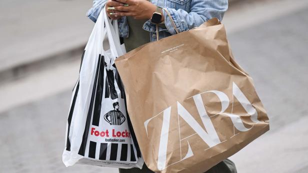 Eine Person trägt Einkaufstüten von Zara und Foot Locker und benutzt ein Smartphone.