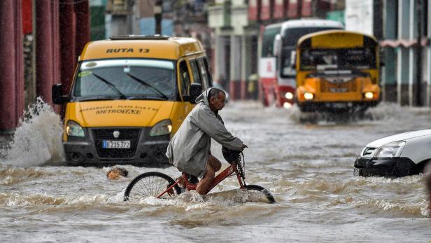 Ein Mann fährt mit dem Fahrrad durch eine überflutete Straße in Kuba, während Busse und Autos durch das Wasser fahren.