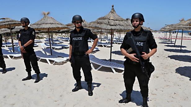 Bewaffnete Polizisten patrouillieren an einem sonnigen Strand mit Sonnenschirmen und Liegestühlen.