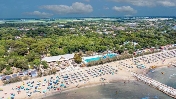 Luftaufnahme eines belebten Strandes mit bunten Sonnenschirmen und einem Schwimmbad im Hintergrund.