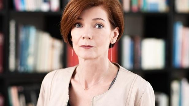 Eine Frau mit roten Haaren und hellem Blazer steht vor einem Bücherregal.