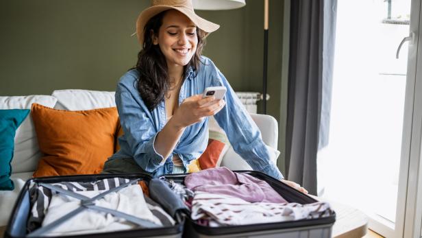 Eine lächelnde Frau mit Hut packt Kleidung in einen offenen Koffer und schaut auf ihr Smartphone.