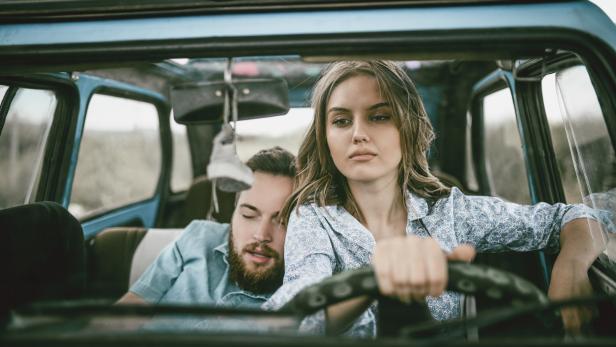 Eine junge Frau fährt einen blauen Oldtimer, während ein Mann auf dem Beifahrersitz schläft.