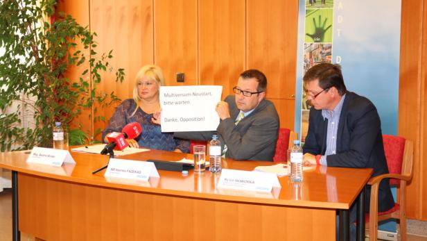 Drei Personen sitzen an einem Tisch, eine hält ein Schild mit der Aufschrift „Multiversum-Neustart, bitte warten. Danke, Opposition!“.