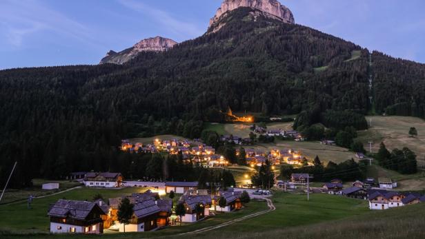 Ein Bergdorf liegt eingebettet in einer grünen Landschaft unter einem hohen Bergmassiv.