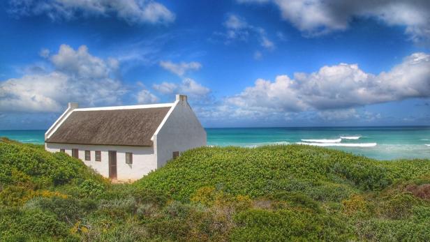 Ein weißes Haus mit Strohdach liegt in Küstennähe hinter grünen Dünen.