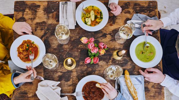 Ein Esstisch mit mehreren Personen, Tellern mit verschiedenen Gerichten, Wein und einem Blumenstrauß.