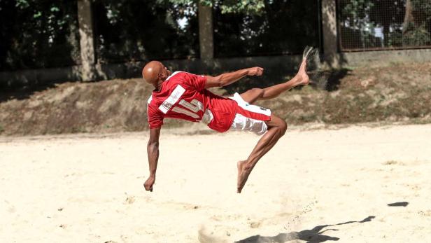 Ein barfüßiger Mann in Sportkleidung springt und tritt einen Ball am Strand.