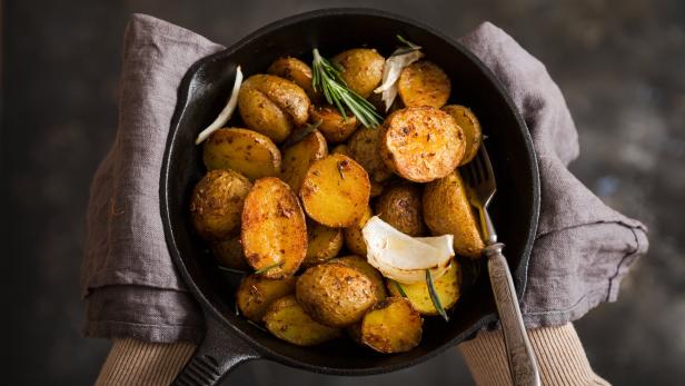 Eine Person hält eine gusseiserne Pfanne mit Bratkartoffeln, Zwiebeln und Rosmarin.