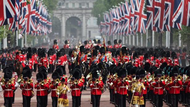 Eine Parade britischer Soldaten in roten Uniformen und Bärenfellmützen marschiert unter Union Jack-Flaggen.
