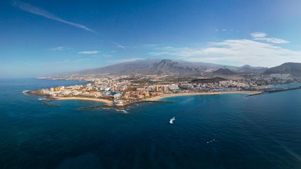 Küstenstadt mit Sandstränden, Hotels und Bergen im Hintergrund, das Meer ist ruhig und ein Boot fährt auf dem Wasser.