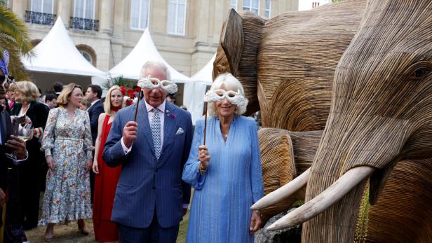 König Charles und Königin Camilla mit Masken vor einer Elefantenskulptur.