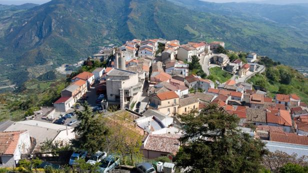 Ein Bergdorf mit roten Ziegeldächern liegt vor einer bewaldeten Hügellandschaft.