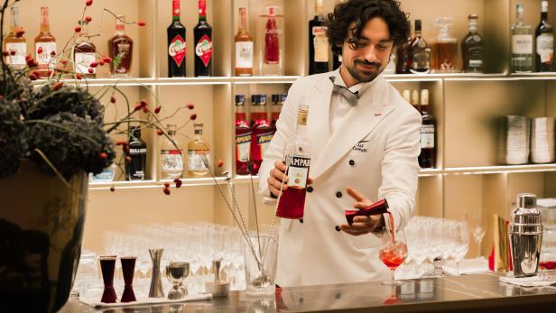 Ein Barkeeper in weißer Jacke mixt einen Cocktail mit Campari an einer Bar.