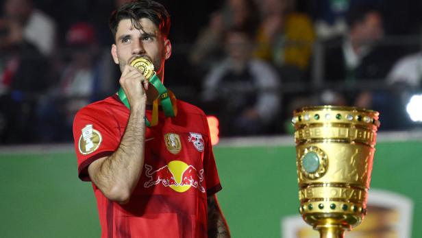 Ein Fußballspieler von RB Leipzig mit einer Goldmedaille und dem DFB-Pokal im Hintergrund.