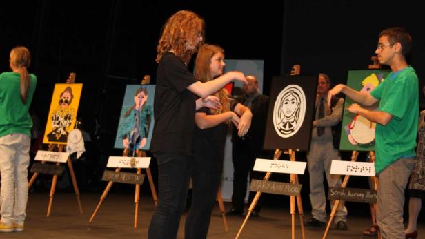 Zwei junge Leute gestikulieren vor einer Reihe von Porträtgemälden auf Staffeleien.