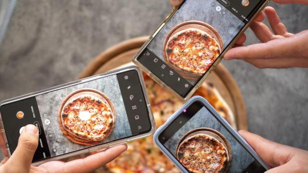 Mehrere Handys fotografieren eine Pizza auf einem Holzteller.