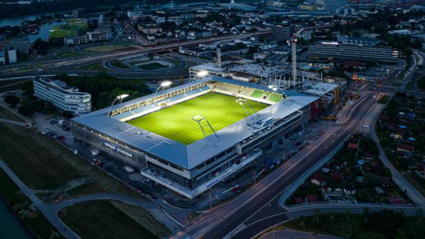 Ein beleuchtetes Fußballstadion mit grünem Spielfeld liegt an einer großen Straßenkreuzung in einer Stadt bei Nacht.