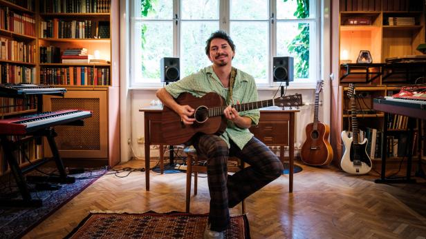 Ein Mann mit Gitarre sitzt in einem Raum voller Bücher und Musikinstrumente.
