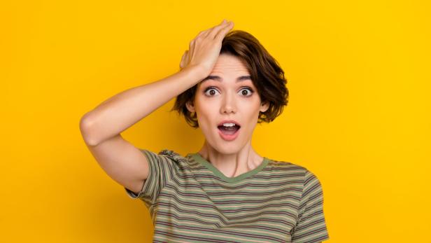 Eine junge Frau mit einem überraschten Gesichtsausdruck hält sich an den Kopf vor einem gelben Hintergrund.