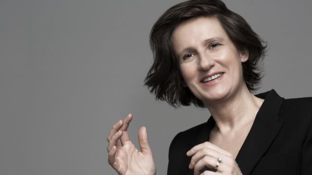 Anna Flipo lächelt in einem schwarzen Blazer vor einem grauen Hintergrund.