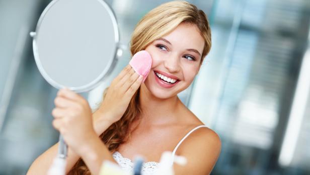 Eine junge Frau reinigt ihr Gesicht mit einem Wattepad vor einem Spiegel.