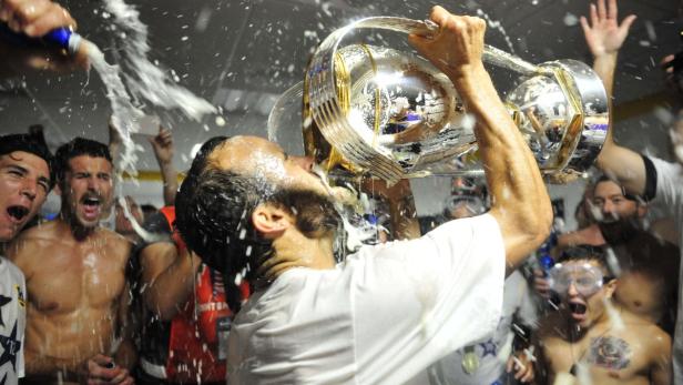 Ein Mann trinkt Champagner aus einem großen silbernen Pokal, umgeben von jubelnden Menschen.