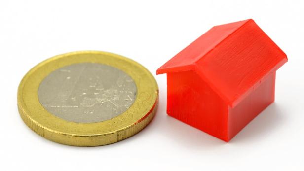 Eine Euro-Münze liegt neben einem kleinen, roten Spielzeughaus.