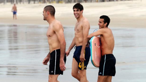 Drei Männer in Badehosen gehen am Strand entlang, einer trägt ein Surfbrett.