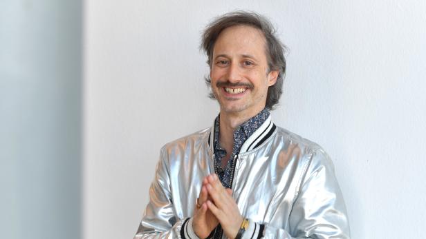 Ein lächelnder Mann mit Schnurrbart und silberner Jacke steht vor einer weißen Wand.