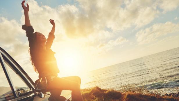 Eine junge Frau sitzt auf einem Auto am Meer und streckt die Arme in die Luft.