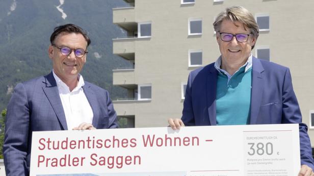 Zwei Männer präsentieren ein Schild für studentisches Wohnen in Pradler Saggen vor einem Gebäude und Bergkulisse.