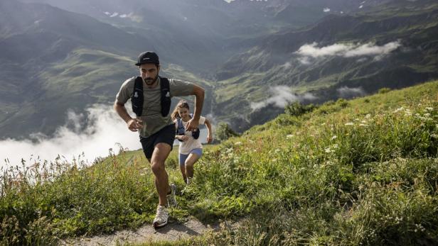 Ein Mann und eine Frau laufen einen steilen, grasbewachsenen Hang vor einer Bergkulisse hinauf.
