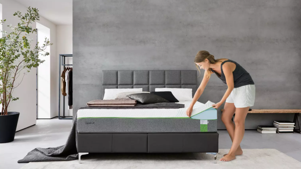 Eine Frau bezieht ein modernes Bett mit grauem Kopfteil in einem minimalistisch eingerichteten Schlafzimmer.