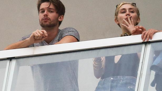 Miley Cyrus raucht auf einem Balkon, neben ihr ein Mann.
